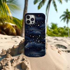 Pode incluir: Uma capa de smartphone com um design de c&eacute;u noturno azul escuro com estrelas douradas, luas crescentes e nuvens. A capa est&aacute; colocada em uma praia arenosa com palmeiras ao fundo.