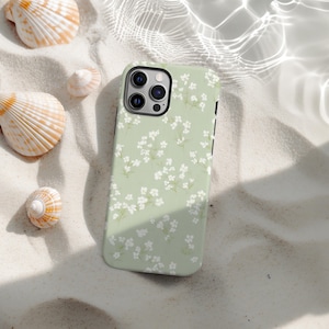 May include: A pale green phone case adorned with a white floral design. The case rests on sand, surrounded by seashells. The phone case features a black camera lens and black side buttons. The case is likely for a smartphone.