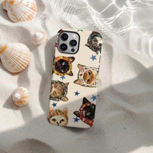 May include: A phone case with a repeating pattern of cats in bonnets and blue stars on a cream background. The case has a camera cutout. Seashells are scattered on a sandy surface around the case.