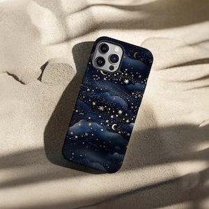Pode incluir: Uma capa de telefone azul escuro com um design celestial de estrelas douradas, luas crescentes e formas de nuvens. A capa est&aacute; em uma superf&iacute;cie arenosa, contrastando com o fundo claro.