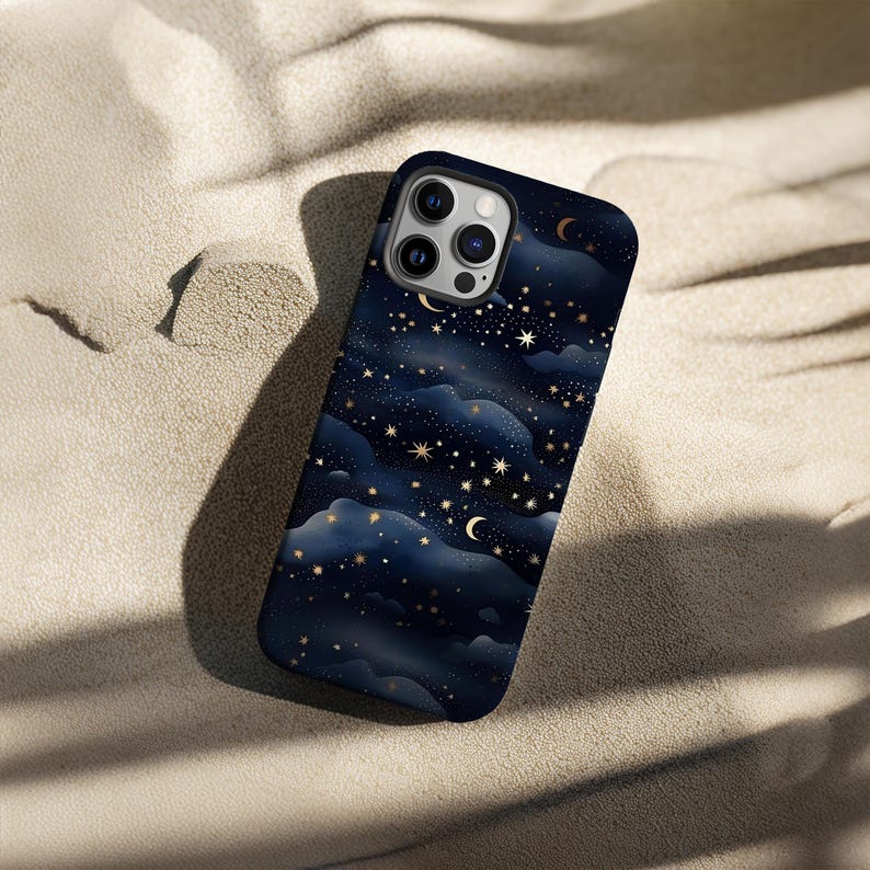 Pode incluir: Uma capa de telefone azul escuro com um design celestial de estrelas douradas, luas crescentes e formas de nuvens. A capa est&aacute; em uma superf&iacute;cie arenosa, contrastando com o fundo claro.