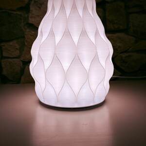 May include: A white, decorative lamp with a unique, textured design. The lamp has a rounded base and a flared top, with a pattern of diamond shapes. The lamp is lit from within, creating a soft glow. The background is a dark stone wall.