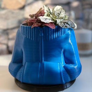 Pode incluir: Um vaso azul em forma de casaco cheio de plantas coloridas. O vaso tem um detalhe de zíper e gola canelada. As plantas têm folhas brancas, rosa e verdes. O vaso assenta numa base circular preta.