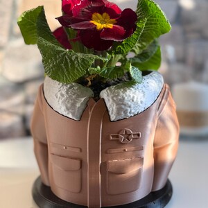 May include: A brown planter shaped like a bomber jacket, with a white fur collar and a star emblem. The planter holds a vibrant plant with dark red flowers and green leaves. The planter sits on a black base.