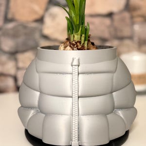Pode incluir: Um vaso de flores prateado em forma de casaco acolchoado, com um fecho central. O vaso contém uma planta em crescimento com folhas verdes. A base é preta. O fundo é uma parede de pedra desfocada.