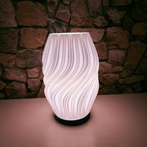 Könnte beinhalten: Eine weiße Tischlampe mit Wirbeldesign und schwarzem Sockel. Der Lampenschirm weist vertikale Linien auf, die ein strukturiertes, wellenartiges Muster erzeugen. Die Lampe ist beleuchtet und wirft ein sanftes Leuchten. Der Hintergrund ist eine Steinmauer.