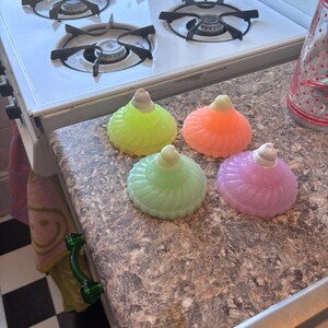 May include: Four glass candle holders in different colors: green, orange, light green, and purple. The candle holders are shaped like a dome with a rounded top and a fluted design. They are sitting on a brown countertop.
