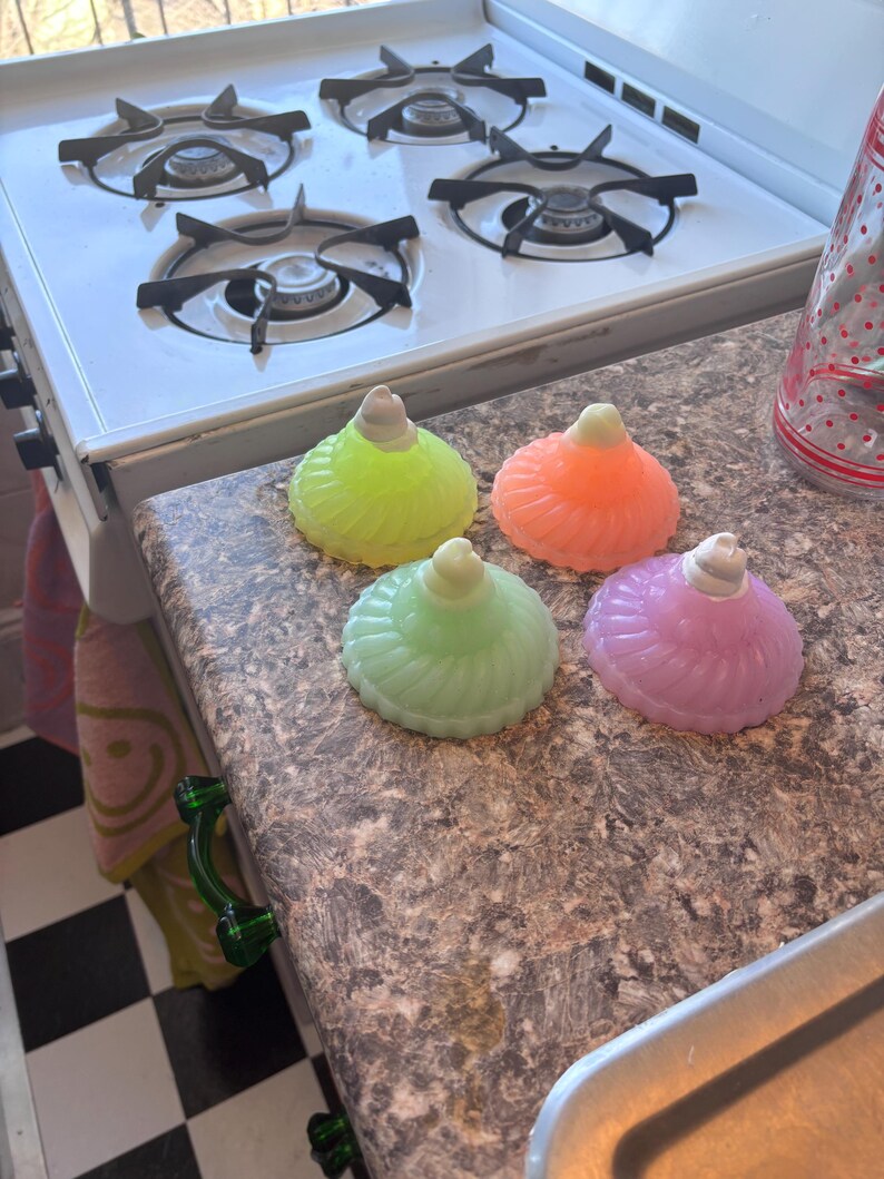 May include: Four glass candle holders in different colors: green, orange, light green, and purple. The candle holders are shaped like a dome with a rounded top and a fluted design. They are sitting on a brown countertop.