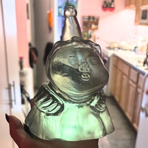 May include: A clear glass figurine of a character with a pointed hat and a surprised expression. The figurine is lit from within, casting a green glow.