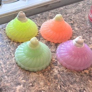 May include: Four colorful glass bottle stoppers in the shape of a ruffled dress. The stoppers are in shades of green, pink, yellow, and orange.