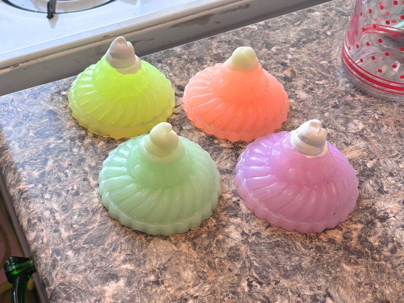 May include: Four colorful glass bottle stoppers in the shape of a ruffled dress. The stoppers are in shades of green, pink, yellow, and orange.