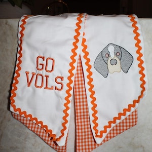 May include: A white fabric scarf featuring orange rickrack trim and an orange gingham underside. One end displays the embroidered text GO VOLS in orange, while the other end features an embroidered gray and white dog head graphic.