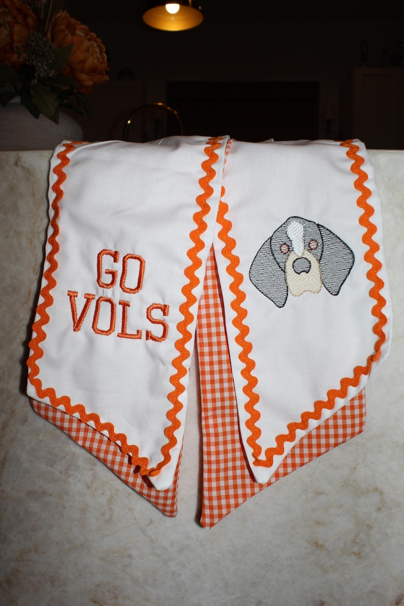 May include: A white fabric scarf featuring orange rickrack trim and an orange gingham underside. One end displays the embroidered text GO VOLS in orange, while the other end features an embroidered gray and white dog head graphic.