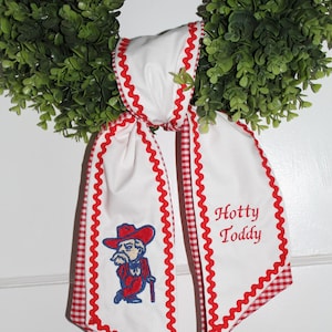 May include: A white fabric ribbon with red gingham trim and red rickrack edging. One side features an embroidered mascot in a red hat and suit with a cane, while the other side displays the embroidered red text Hotty Toddy on a green wreath.