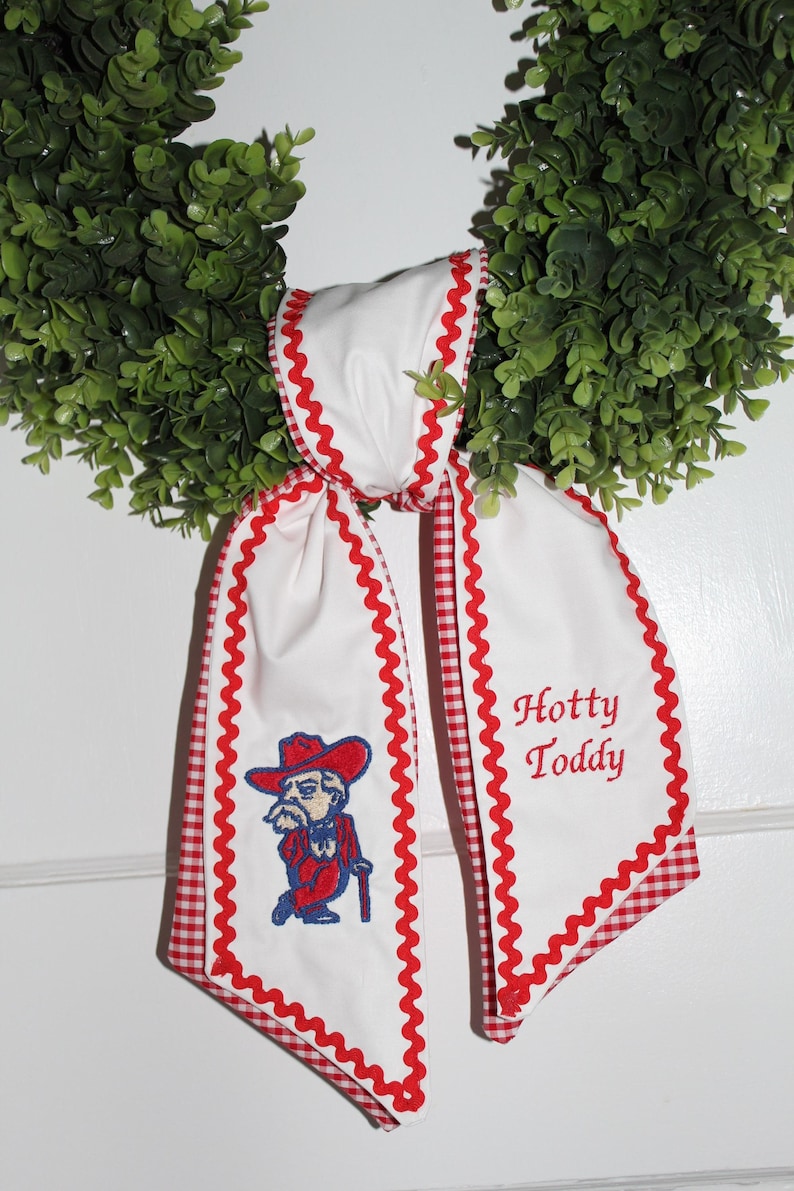 May include: A white fabric ribbon with red gingham trim and red rickrack edging. One side features an embroidered mascot in a red hat and suit with a cane, while the other side displays the embroidered red text Hotty Toddy on a green wreath.
