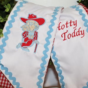 May include: A white fabric banner featuring light blue rickrack trim along the edges. The banner displays an embroidered red cowboy hat-wearing figure with a cane and the red embroidered text Hotty Toddy. The reverse side shows a red and white gingham pattern.