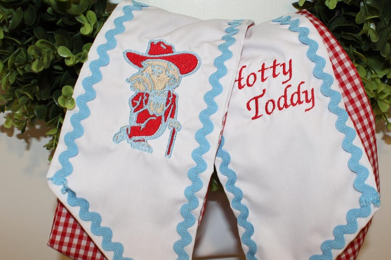 May include: A white fabric banner featuring light blue rickrack trim along the edges. The banner displays an embroidered red cowboy hat-wearing figure with a cane and the red embroidered text Hotty Toddy. The reverse side shows a red and white gingham pattern.