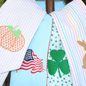 May include: A collection of decorative kitchen towels with seasonal and holiday-themed embroidery. Designs include a pumpkin, American flag, shamrock, and a bunny with a yellow bow. The towels have striped and patterned backgrounds.