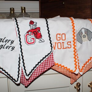 May include: Four white kitchen towels with decorative trim. Two towels feature black rickrack trim and red gingham fabric, one with black embroidered text glory glory and one with a bulldog mascot and letter G. Two towels feature orange rickrack trim and orange gingham fabric, one with orange embroidered text GO VOLS and one with a dog face graphic.