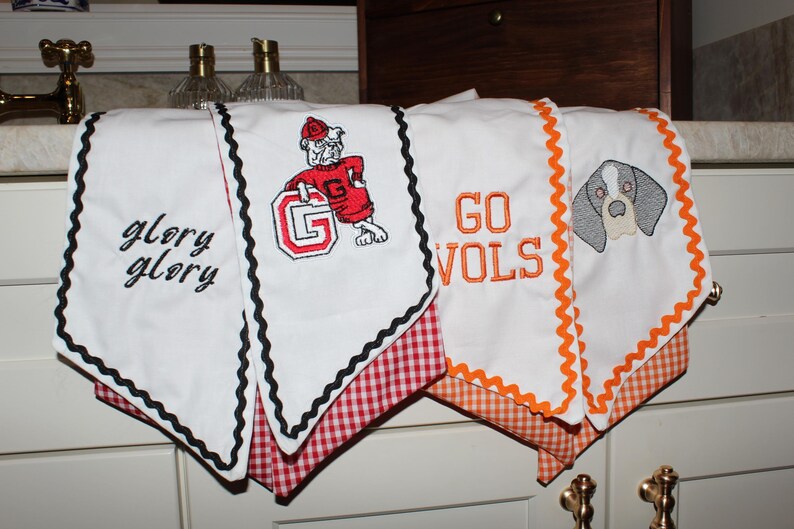 May include: Four white kitchen towels with decorative trim. Two towels feature black rickrack trim and red gingham fabric, one with black embroidered text glory glory and one with a bulldog mascot and letter G. Two towels feature orange rickrack trim and orange gingham fabric, one with orange embroidered text GO VOLS and one with a dog face graphic.