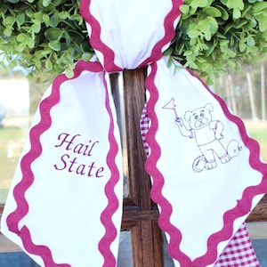 May include: A white fabric door hanger featuring a maroon rickrack trim and a red and white gingham backing. The left side displays the maroon text Hail State, while the right side features a maroon embroidered graphic of a bulldog holding a flag.