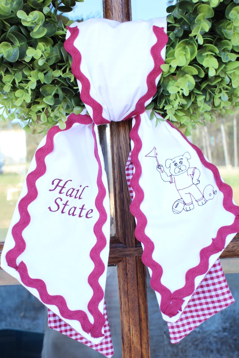 May include: A white fabric door hanger featuring a maroon rickrack trim and a red and white gingham backing. The left side displays the maroon text Hail State, while the right side features a maroon embroidered graphic of a bulldog holding a flag.