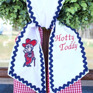May include: A white fabric door hanger featuring a red and white gingham backing, trimmed with navy blue rickrack. The front displays an embroidered mascot in a red suit and hat, alongside the red embroidered text Hotty Toddy.
