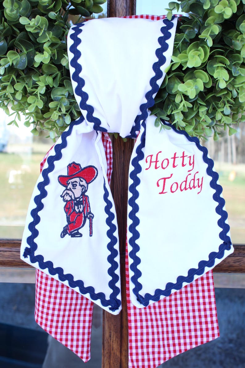 May include: A white fabric door hanger featuring a red and white gingham backing, trimmed with navy blue rickrack. The front displays an embroidered mascot in a red suit and hat, alongside the red embroidered text Hotty Toddy.