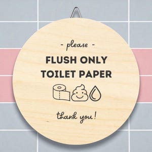 Puede incluir: Un letrero redondo de madera con un gancho de metal. El letrero tiene el texto "- please - FLUSH ONLY TOILET PAPER thank you!" con ilustraciones de papel higiénico, un emoji de caca sonriente y una gota de agua.