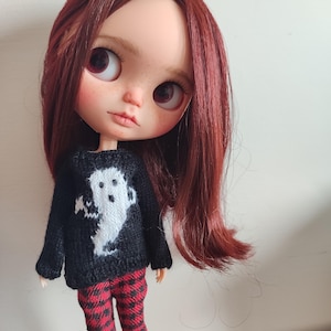 May include: A doll with long, auburn hair wearing a black sweater with a white ghost design and red and black plaid pants.