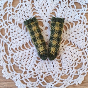 May include: Two miniature green and yellow patterned knitted socks rest on a white, openwork, crocheted doily. The doily is circular with a scalloped edge. The background is a light brown wooden surface.