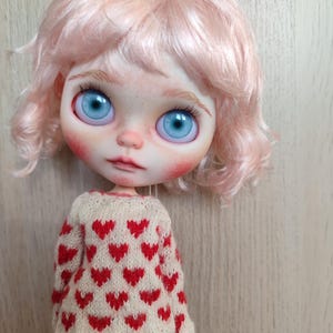 May include: A doll with pink curly hair wearing a cream sweater with red hearts and a pair of red shorts.