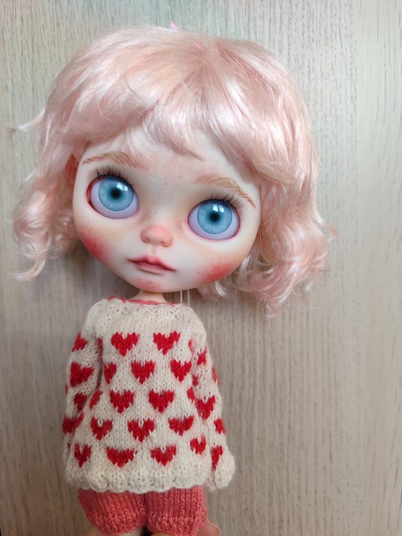 May include: A doll with pink curly hair wearing a cream sweater with red hearts and a pair of red shorts.