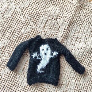 May include: Black knitted sweater with a white ghost design. The ghost has a face and outstretched arms.