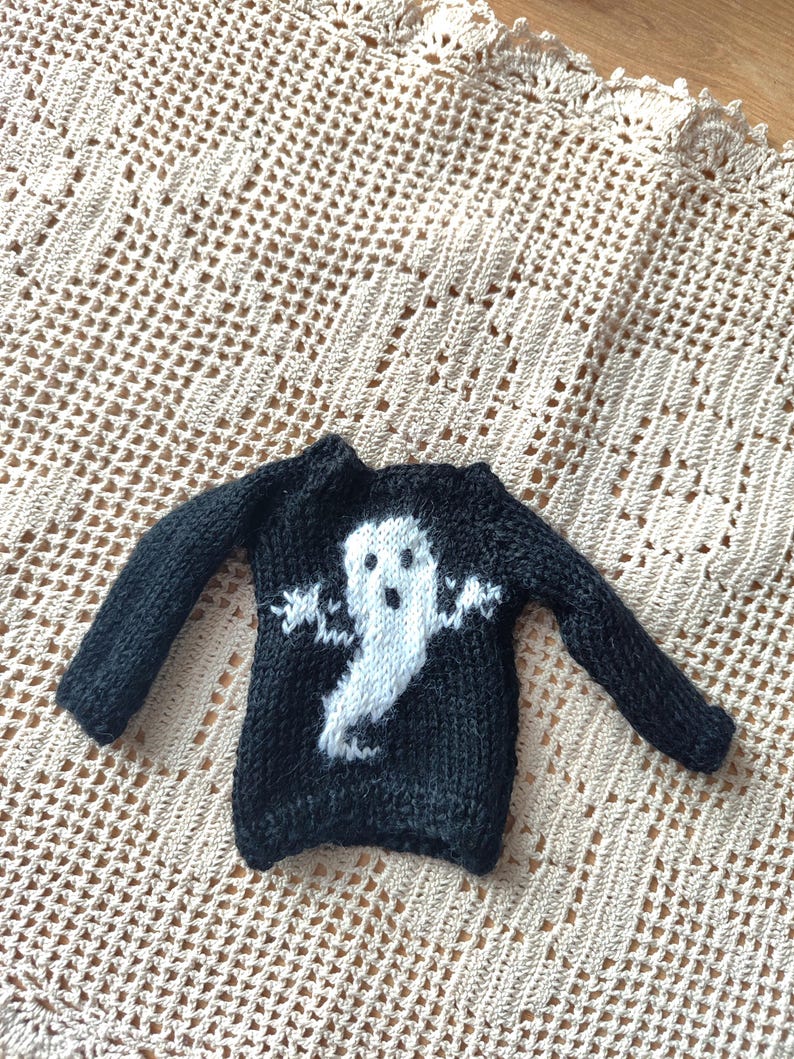 May include: Black knitted sweater with a white ghost design. The ghost has a face and outstretched arms.