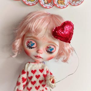 May include: A pink haired doll with a red heart balloon. The doll is wearing a white sweater with red hearts and pink shorts. The doll has blue eyes with a bird design.