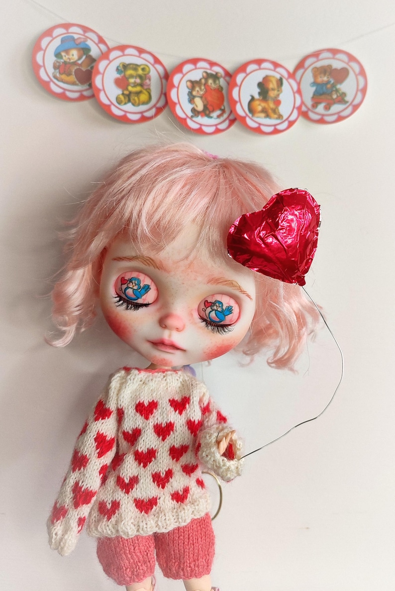 May include: A pink haired doll with a red heart balloon. The doll is wearing a white sweater with red hearts and pink shorts. The doll has blue eyes with a bird design.