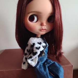 May include: A doll with long, dark red hair wearing a white sweater with black hearts and blue overalls.