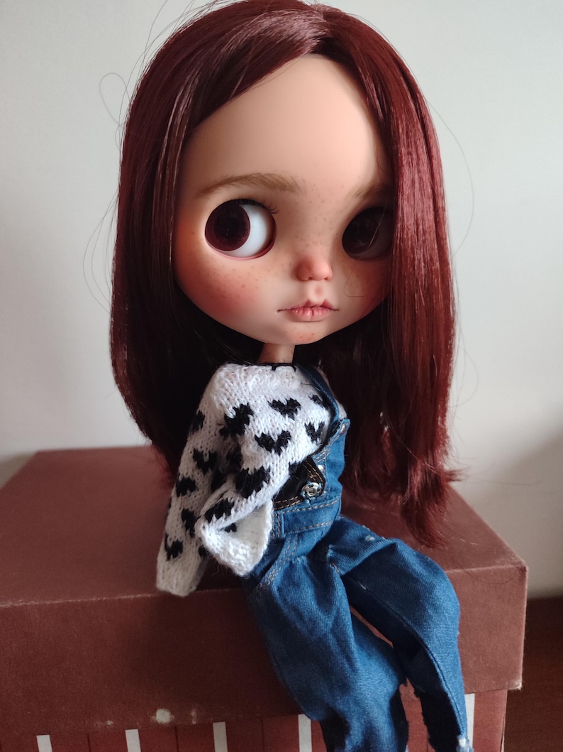 May include: A doll with long, dark red hair wearing a white sweater with black hearts and blue overalls.