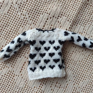 May include: A white knitted sweater with a black heart pattern. The sweater has long sleeves and a round neckline.