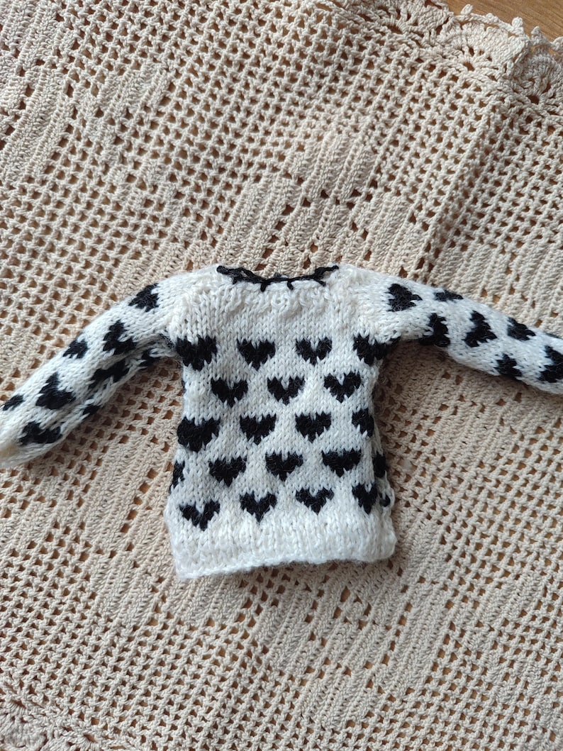 May include: A white knitted sweater with a black heart pattern. The sweater has long sleeves and a round neckline.