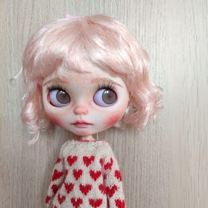 May include: A doll with pink curly hair wearing a white sweater with red hearts. The doll has brown eyes and rosy cheeks.