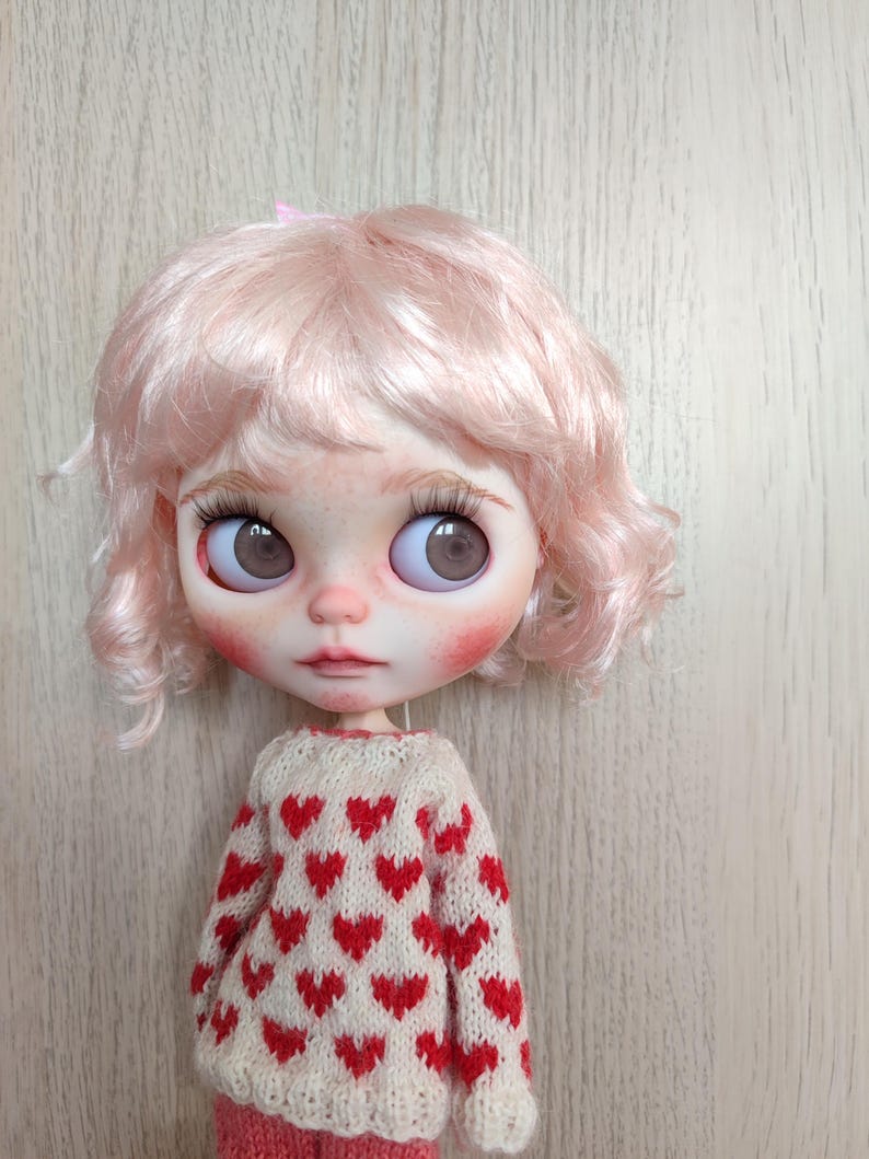 May include: A doll with pink curly hair wearing a white sweater with red hearts. The doll has brown eyes and rosy cheeks.