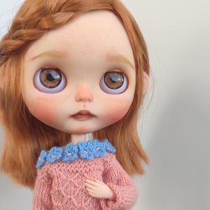 Può includere: Bambola con capelli rossi acconciati a treccia, indossa un maglione rosa con colletto floreale blu e pantaloncini blu. La bambola ha grandi occhi marroni e guance rosee. L'outfit della bambola è lavorato a maglia.