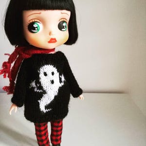 May include: A black and white doll with large green eyes wearing a black sweater with a white ghost design, red and black plaid pants, and black shoes.