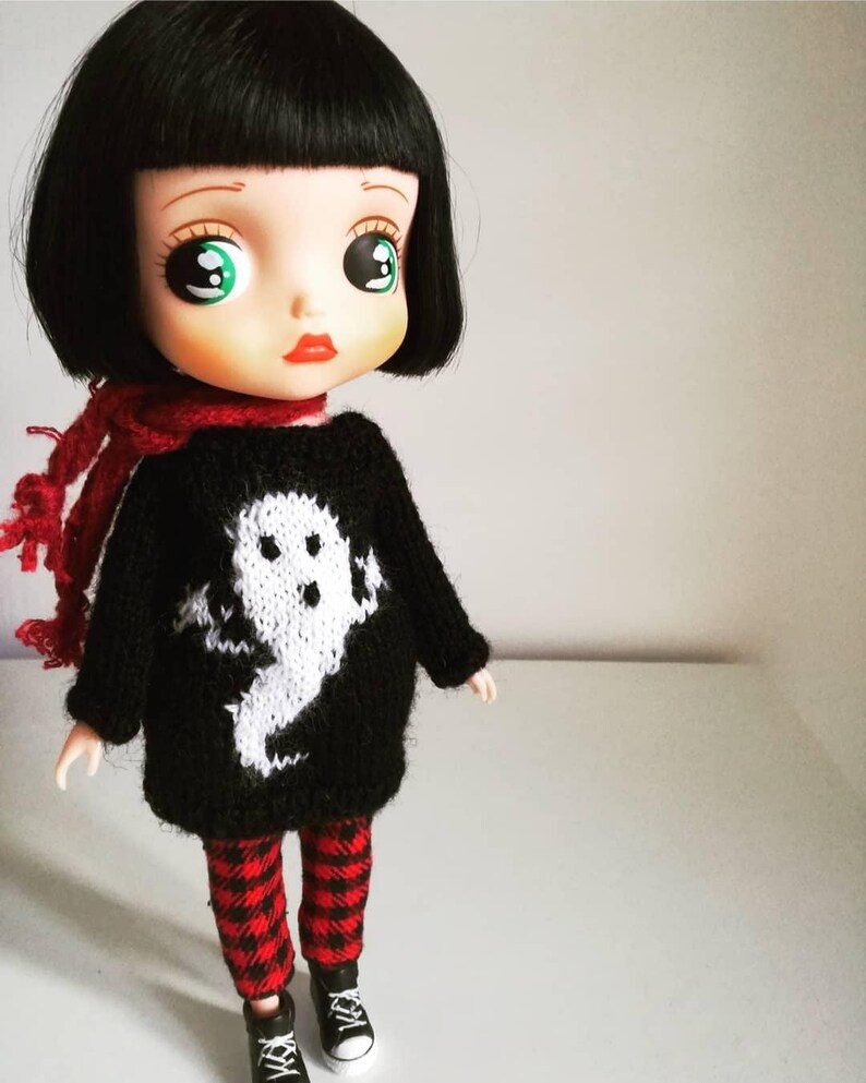 May include: A black and white doll with large green eyes wearing a black sweater with a white ghost design, red and black plaid pants, and black shoes.