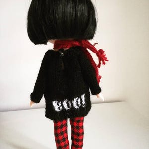 May include: A black knitted sweater with a white "BOO!" design on the back, worn by a doll with black hair, red and black plaid pants, and a red scarf.