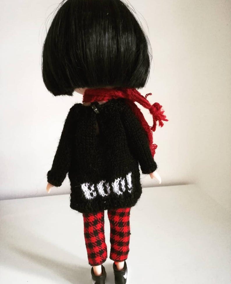 May include: A black knitted sweater with a white "BOO!" design on the back, worn by a doll with black hair, red and black plaid pants, and a red scarf.