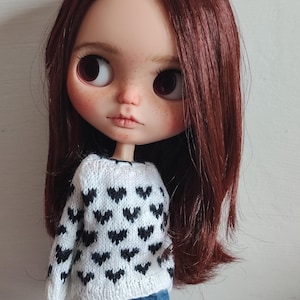 May include: A doll with long, dark red hair wearing a white sweater with a black heart pattern. The doll is wearing blue jeans and has a neutral expression.