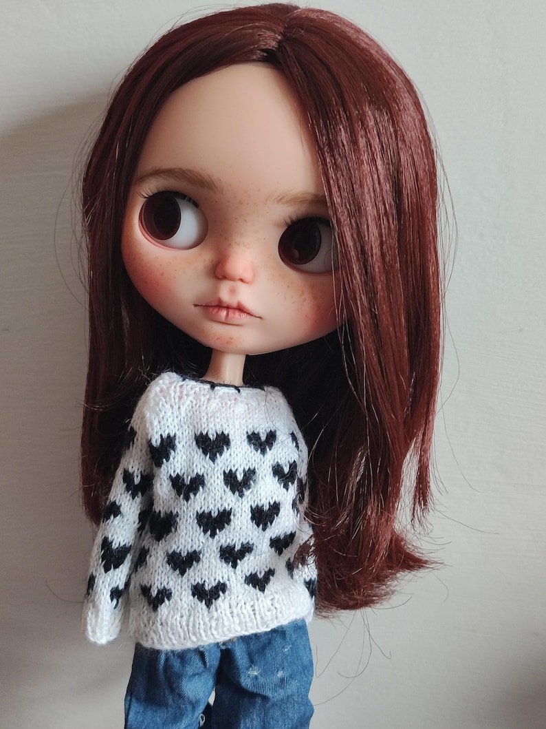 May include: A doll with long, dark red hair wearing a white sweater with a black heart pattern. The doll is wearing blue jeans and has a neutral expression.